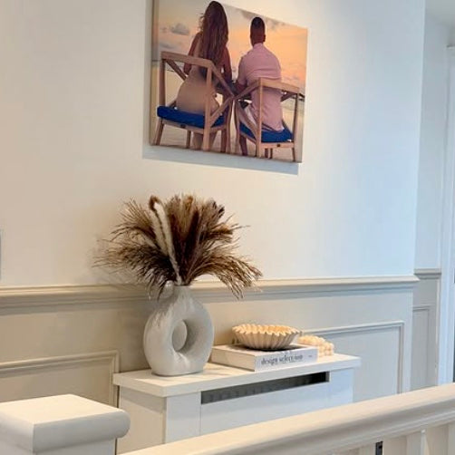 Decorative setup with a nordic vase, dried pampas and books on a white surface, with a painting of a couple in the background.
