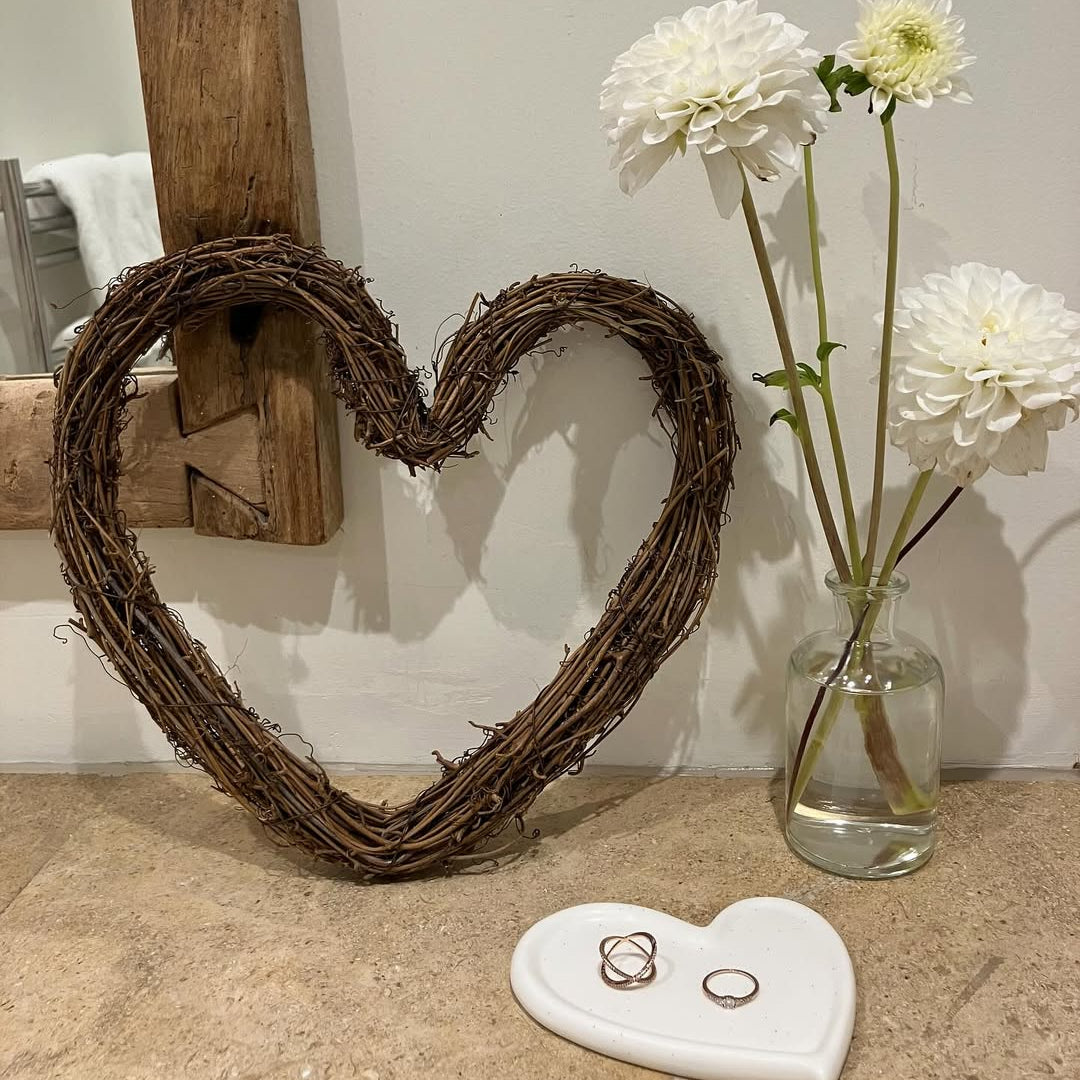 Heart-shaped wreath, white flowers in a vase, and rings on a stone surface.