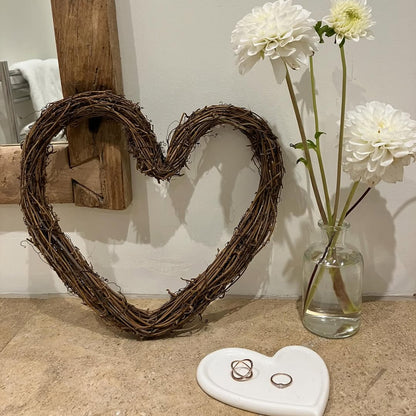 Heart-shaped wreath, white flowers in a vase, and rings on a stone surface.