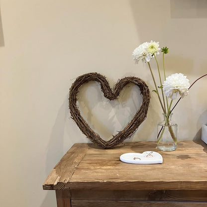 Wooden table with a heart-shaped wreath and vase of flowers against a plain wall.