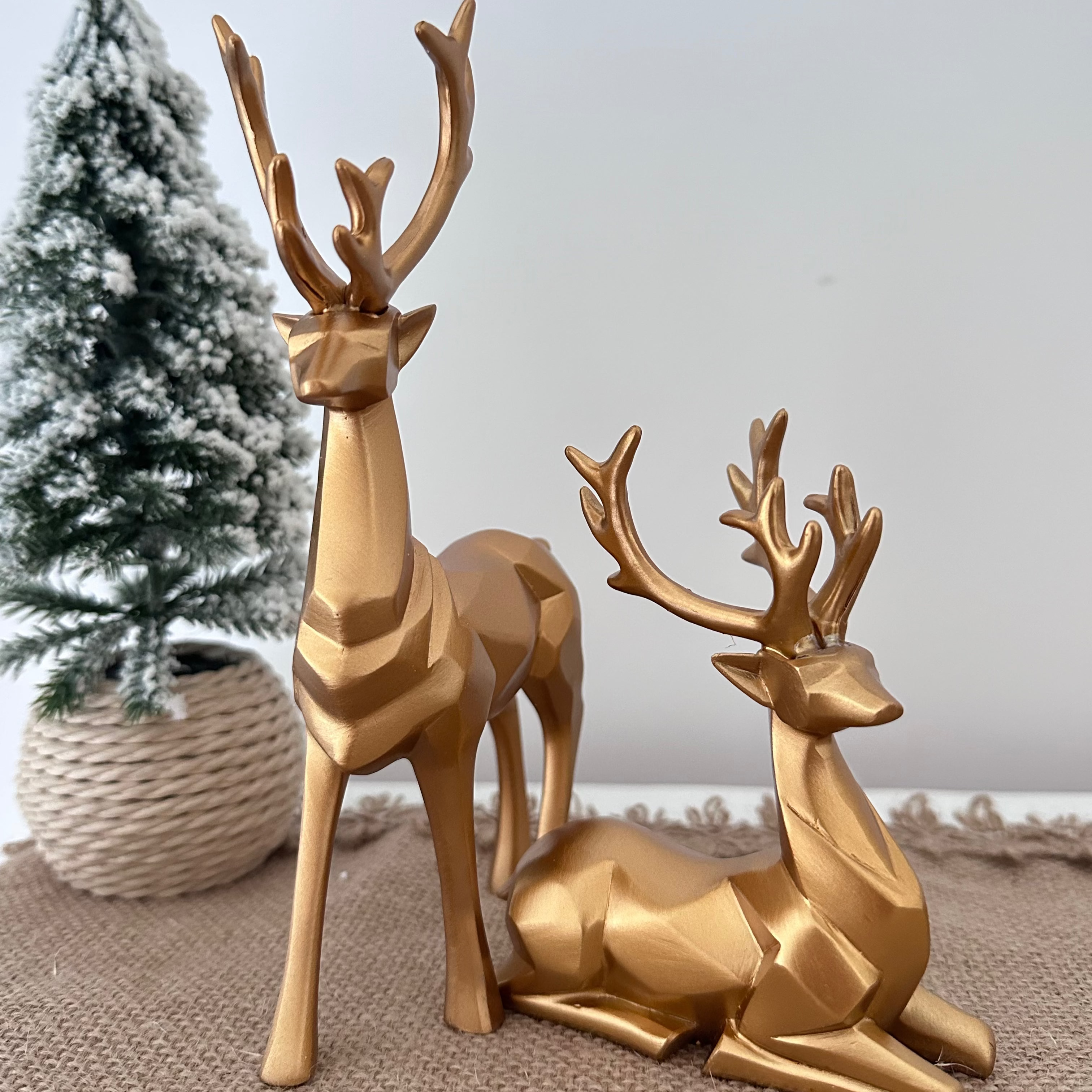 Two gold deer figurines on a textured surface with a small Christmas tree in the background.