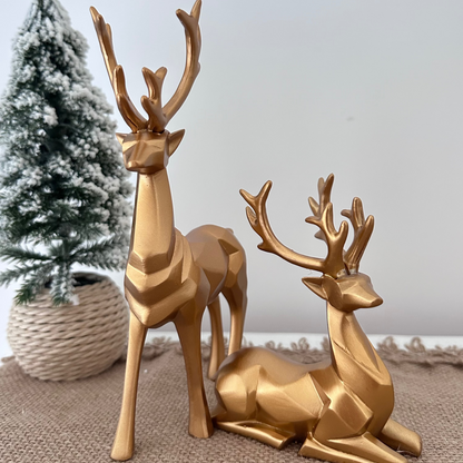 Two gold deer figurines on a textured surface with a small Christmas tree in the background.