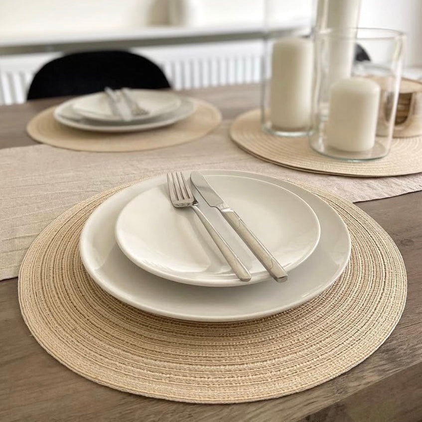Dining table setting with plates, cutlery, and linen, woven placemats on a wooden table.