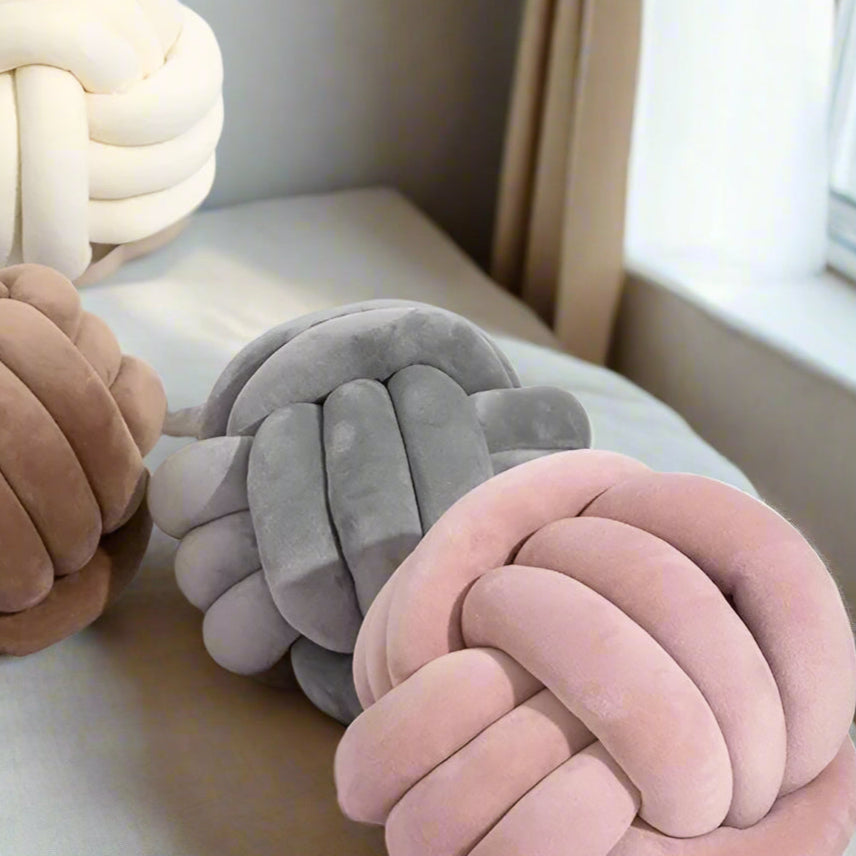 Three knotted cushions in coffee, light grey and pink on a light surface with a neutral background.