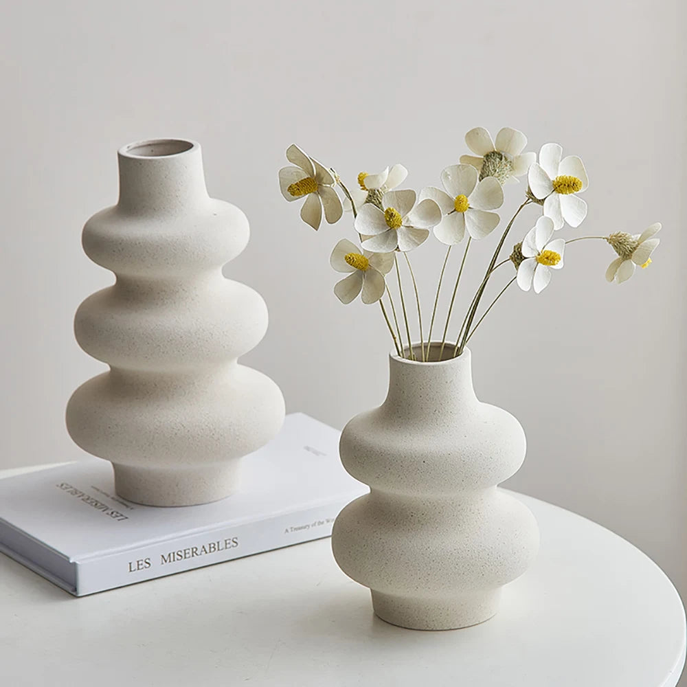 Two white  ceramic vases with a wavy design, one smaller than the other, placed on a surface with a book and flowers in the smaller vase.