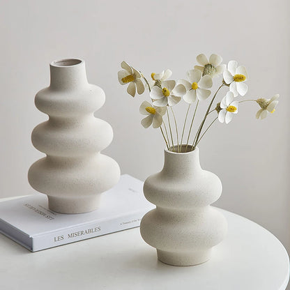 Two white  ceramic vases with a wavy design, one smaller than the other, placed on a surface with a book and flowers in the smaller vase.