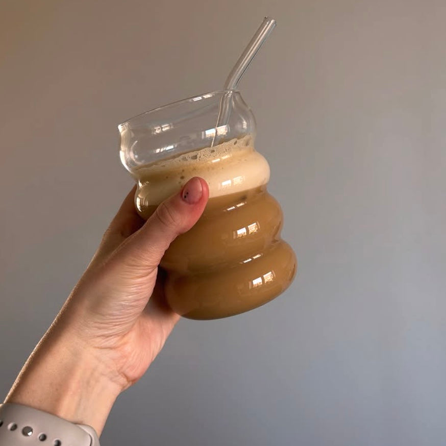 Hand holding a wavy, clear glass with a layered drink and straw, against a plain background