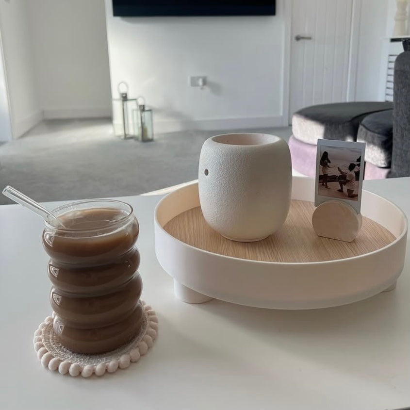 Wavy, clear glass tumbler containing glass straw. The glass is filled with iced coffee. Next to the drink is a round wooden tray with a photo frame in a modern living room.
