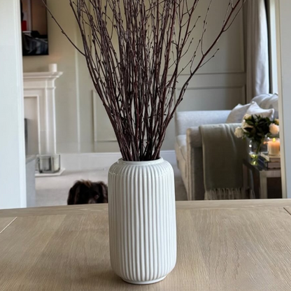 White vase with dried branches on a wooden floor, reflected in a mirror.