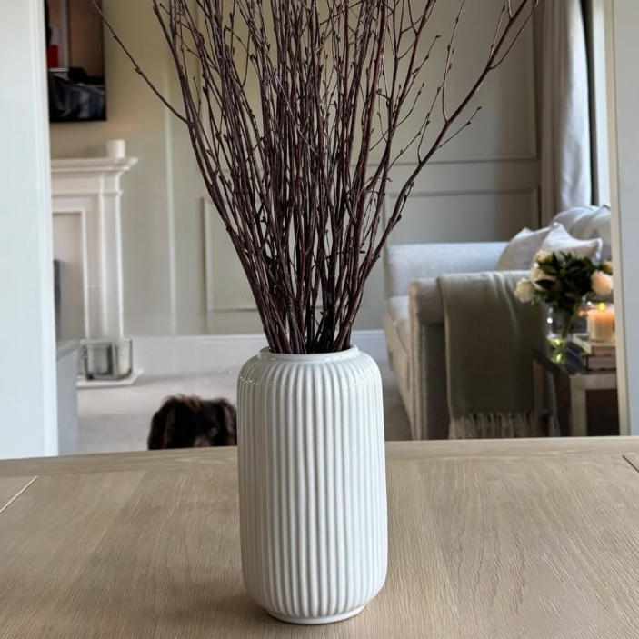 White vase with dried branches on a wooden floor, reflected in a mirror.