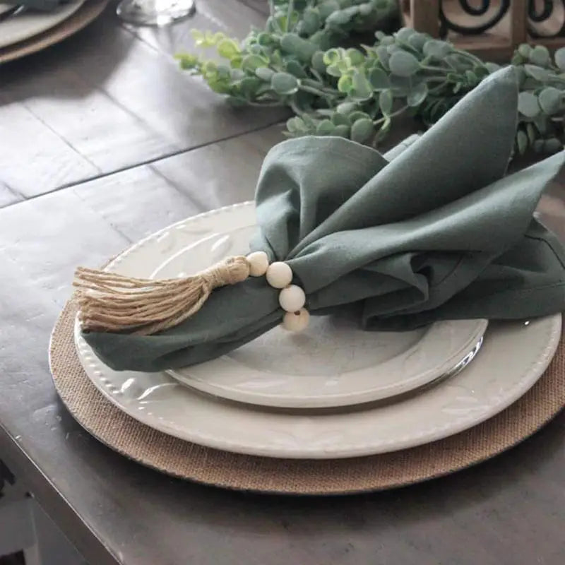 Hand-woven, natural finish, wooden bead napkin rings with jute tassels. They are styled on a dining table. 