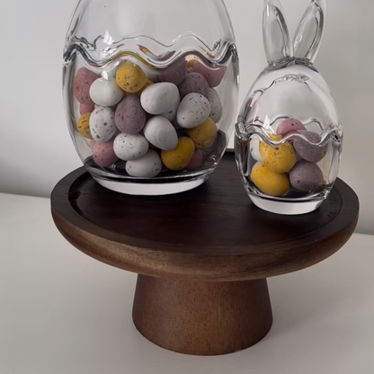 A natural wooded cake stand. On top are two bunny shaped, clear glass jars. The jars are filled with mini Easter eggs. 