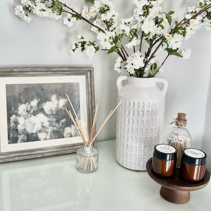 Decorative setup with a vase of white flowers, candles, and a diffuser on a shelf. A small wooden cake stand has been repurposed to support scented decorative candles. 