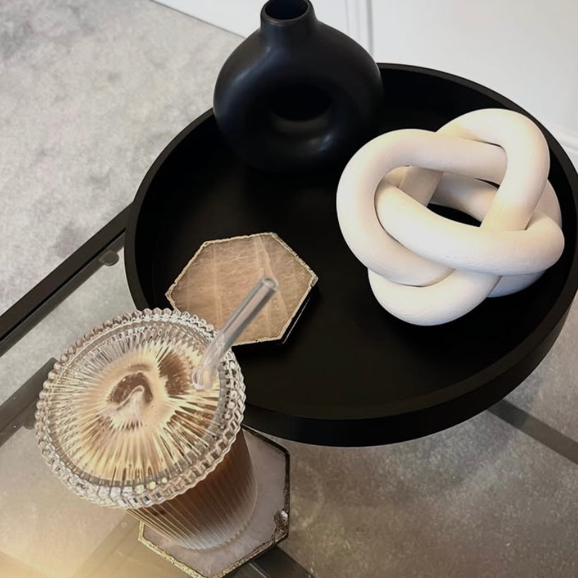 Black round table with decorative items including a glass with a straw, black vase, and white knot sculpture.